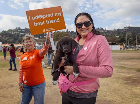 Person holding a puppy at the Los Angeles Super Adoption while another person behind them holds a sign that says I adopted my best friend